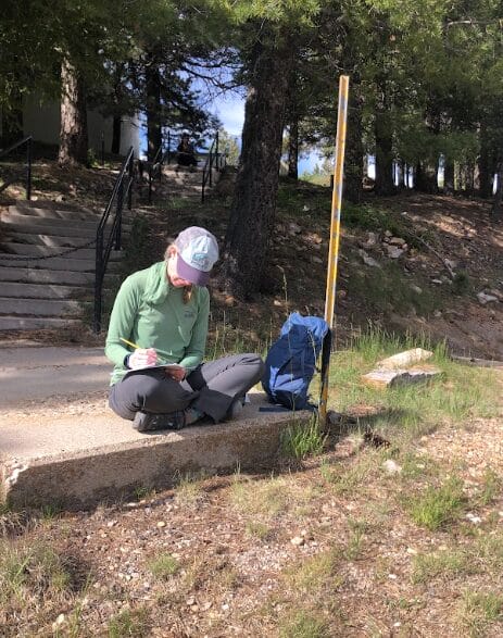 Student sitting cross-legged on sidewalk near forested path, writing notes in a notebook.